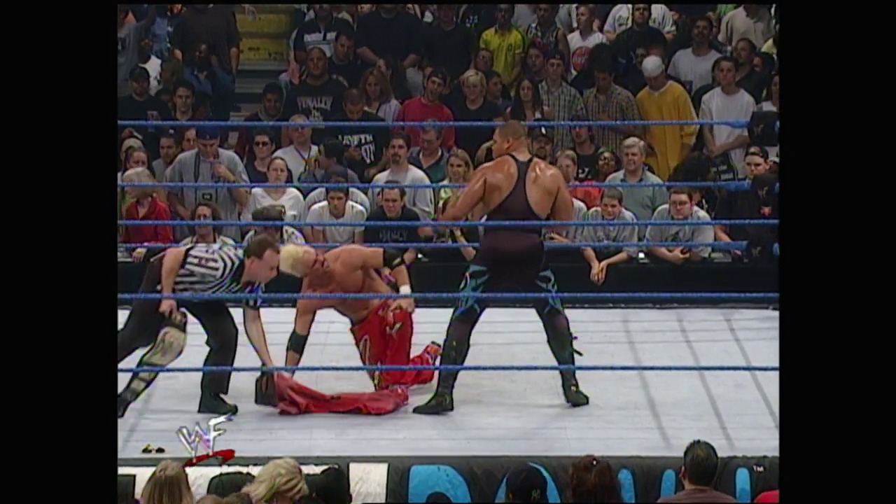 A frame from WWE SmackDown (June 15, 2000) at 38:55. A WWE wrestler in red lies on the canvas while two opponents stand over him in the ring, surrounded by a cheering crowd and blue-roped barriers with the WWF logo visible.