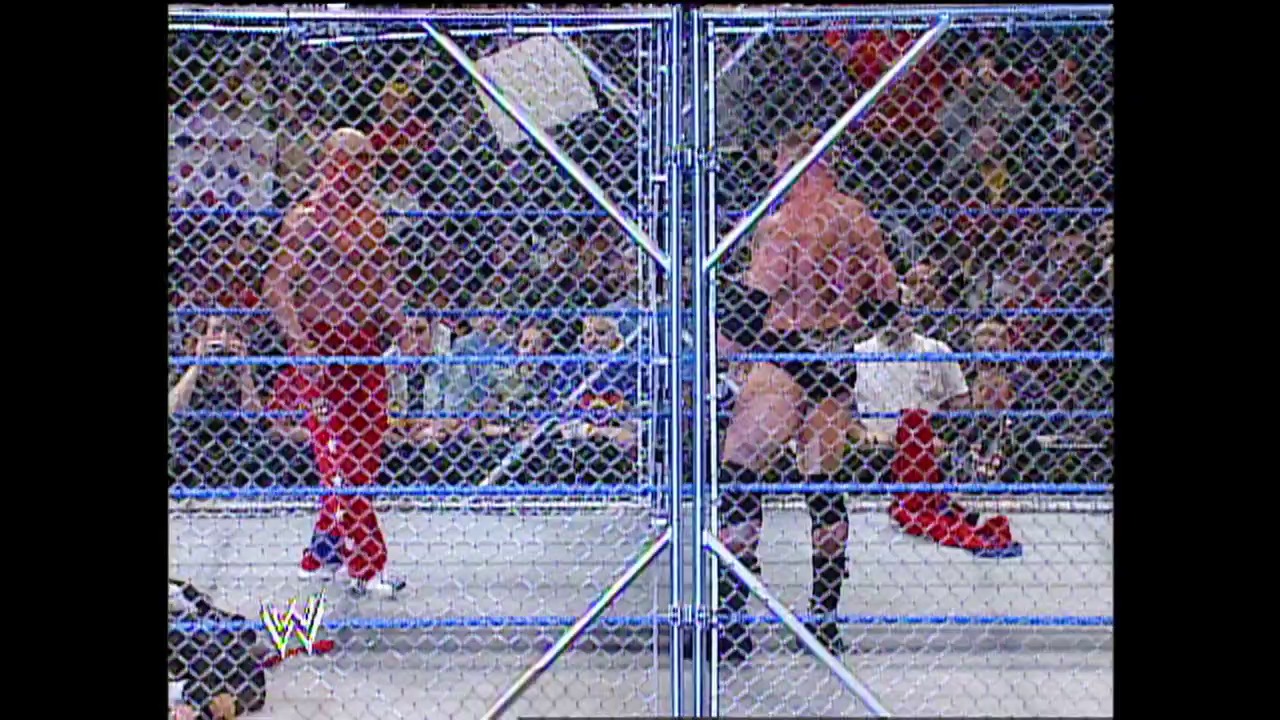 A frame from WWE SmackDown (March 6, 2003) at 1:26:39. A Steel Cage match in progress inside a WWE wrestling ring, with wrestlers visible inside and outside the cage structure. The crowd fills the arena stands in the background, and the WWE logo appears in the bottom left corner of the broadcast.