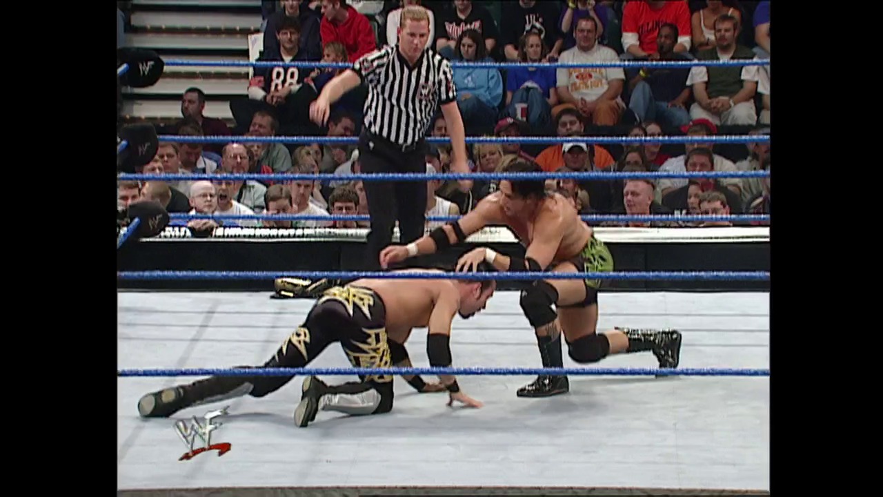 A frame from WWE SmackDown (October 11, 2001) at 39:22. A WWE wrestler in black and gold attire executes a move on the mat while an opponent lies prone, with a referee in striped shirt overseeing the action. The blue-roped ring is surrounded by a packed crowd in the background.