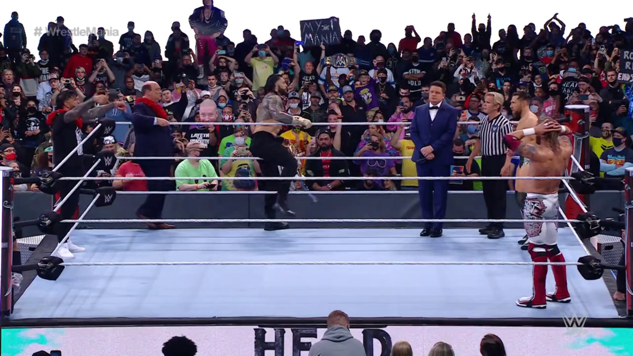 A frame from WWE WrestleMania 37 - Night 2 (2021) at 2:44:36. A WWE wrestling match is in progress with two wrestlers in the ring, a referee in striped shirt, and a man in a tuxedo at ringside. The crowd fills the stadium in the background, with fans holding signs and cheering enthusiastically.