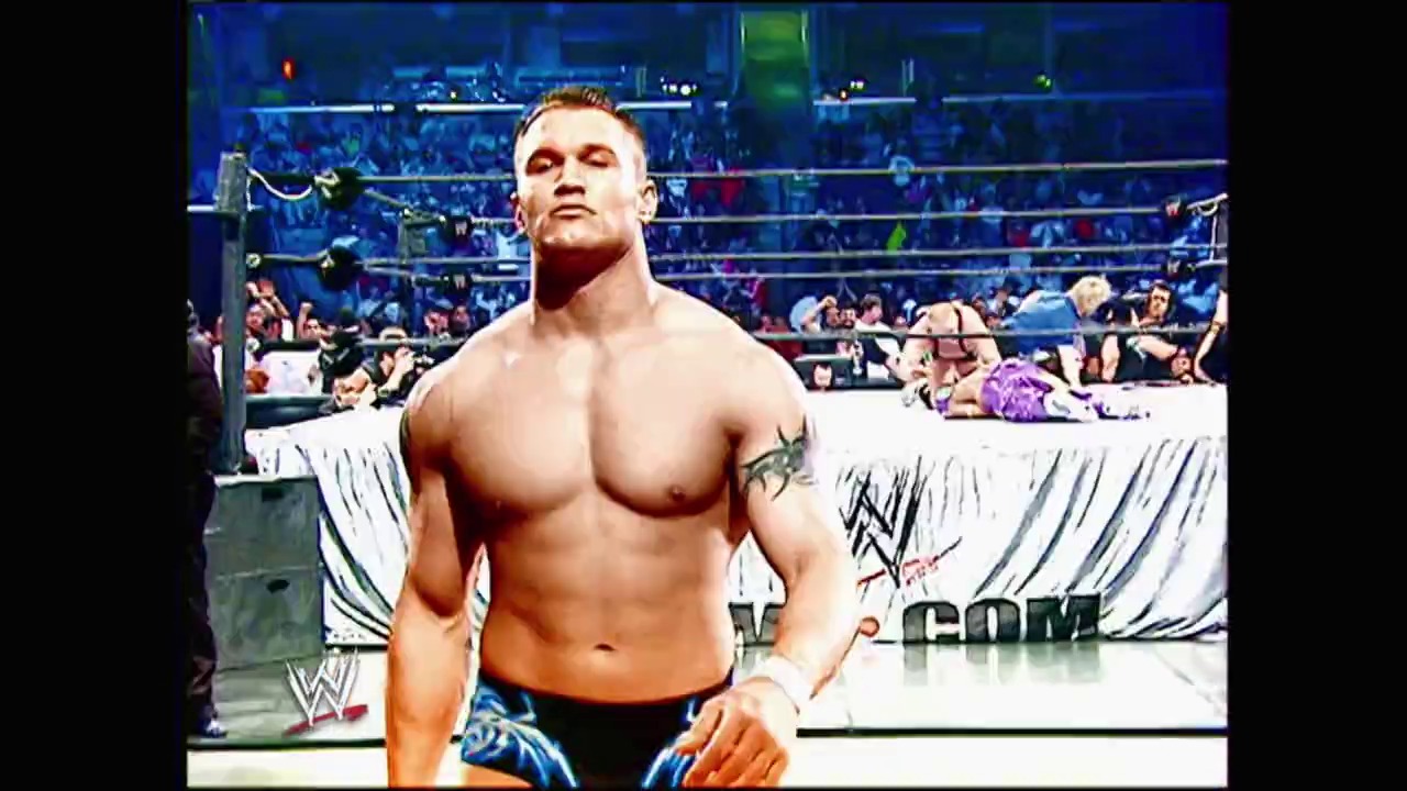 A frame from WrestleMania 22 (2006) at 2:46:13. A muscular wrestler stands in the WWE ring at WrestleMania 22, shirtless and facing the crowd. The arena is filled with spectators in the background, with WWE branding visible on the ring skirts and signage.
