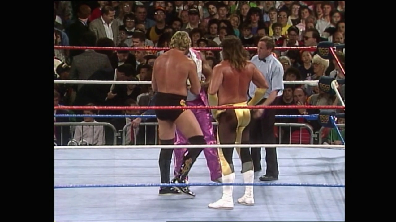 A frame from WrestleMania V (1989) at 29:33. Two shirtless wrestlers face off in a WWE ring surrounded by a packed crowd. A referee in a light blue shirt stands nearby as the wrestlers, one in purple and gold tights and another in black, prepare to engage.