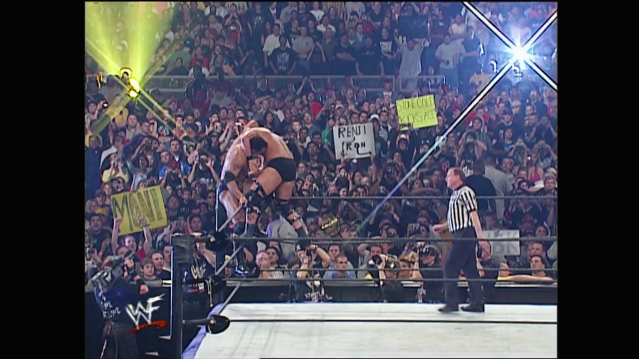 A frame from WrestleMania X-Seven (2001) at 3:12:26. A wrestler performs a move in the ring during WrestleMania X-Seven, with a packed arena crowd visible in the background. Bright stage lighting illuminates the scene, while a referee in striped shirt oversees the match, and spectators hold signs throughout the venue.