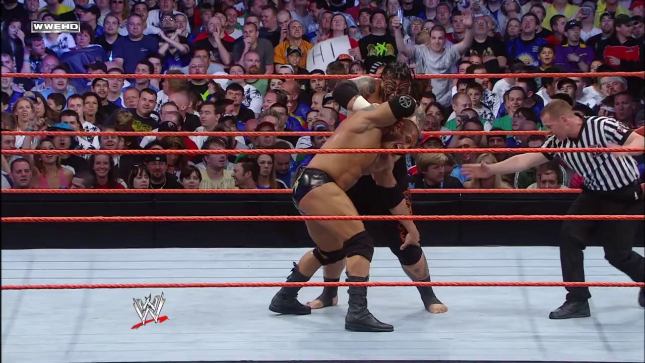 A frame from WrestleMania XXIV (2008) at 1:05:19. A wrestler in black attire executes a move in a WWE ring surrounded by enthusiastic fans. A referee in striped shirt stands nearby as the crowd cheers behind the red ropes, with the WWE logo visible on the mat.