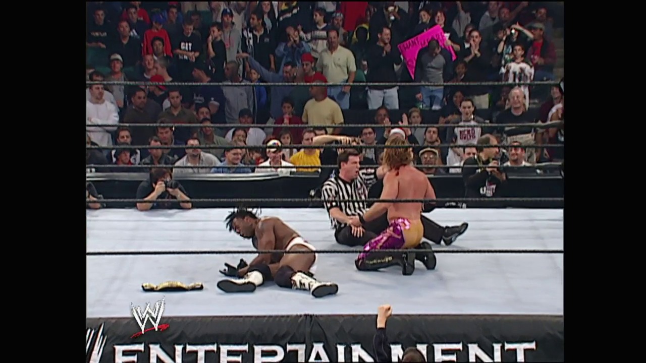 A frame from WWE Armageddon (2002) at 23:59. A WWE wrestling match in progress, with one wrestler down on the mat while another sits nearby as the referee stands between them. The crowd fills the arena stands in the background, with the WWE logo and "Entertainment" text visible at the bottom of the screen.