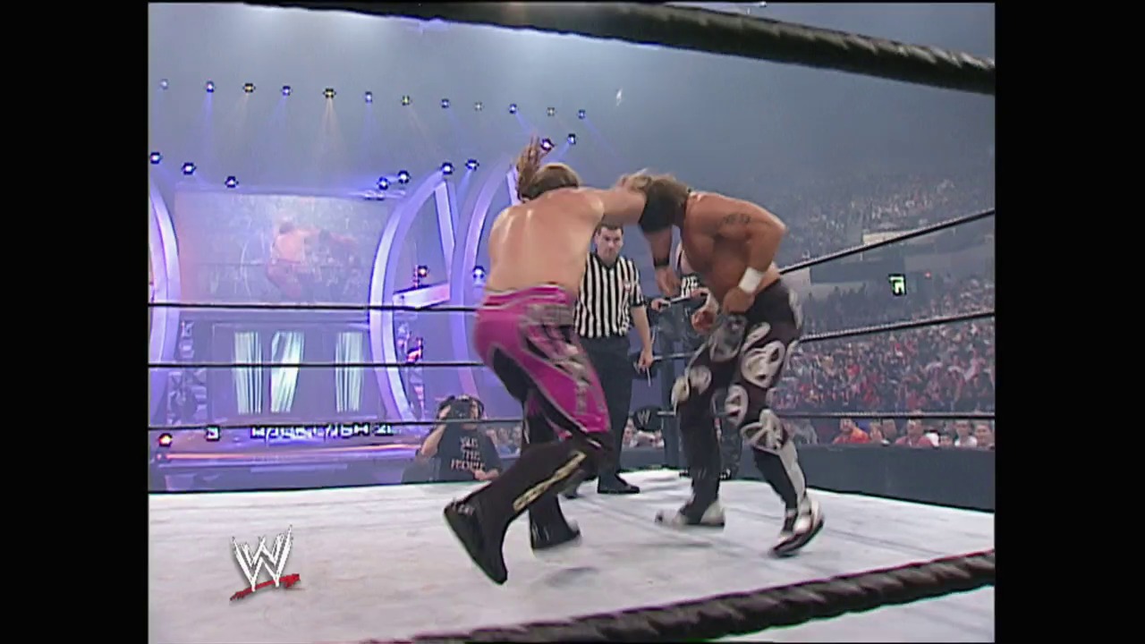 A frame from WWE Backlash (2003) at 1:55:41. A shirtless wrestler in pink/magenta pants delivers a move to an opponent in dark attire inside a WWE wrestling ring, with a referee in striped shirt officiating. The arena is packed with spectators in the background, and purple lighting illuminates the ring structure.