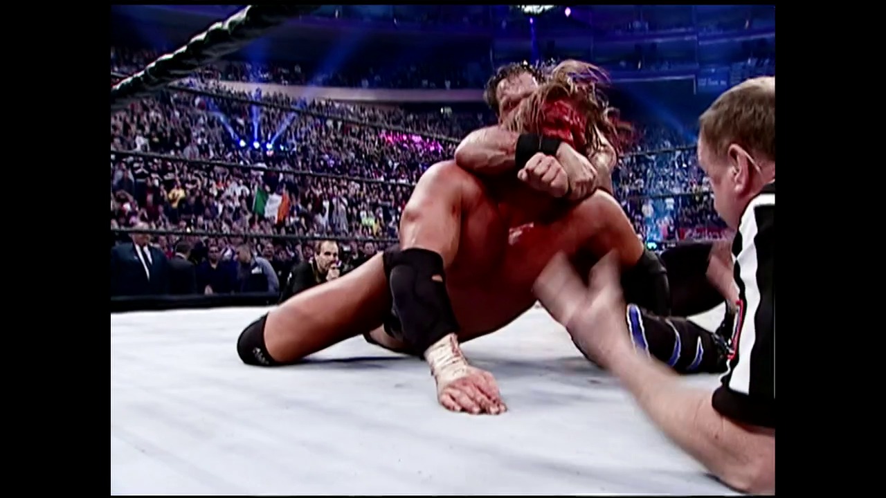 A frame from WWE Backlash (2004) at 0:15. # WWE Backlash 2004

Two wrestlers grapple on the mat inside a packed stadium arena, with one competitor appearing to be in a dominant position. The crowd fills the bleachers in the background under bright blue lighting, while an official monitors the action from ringside.