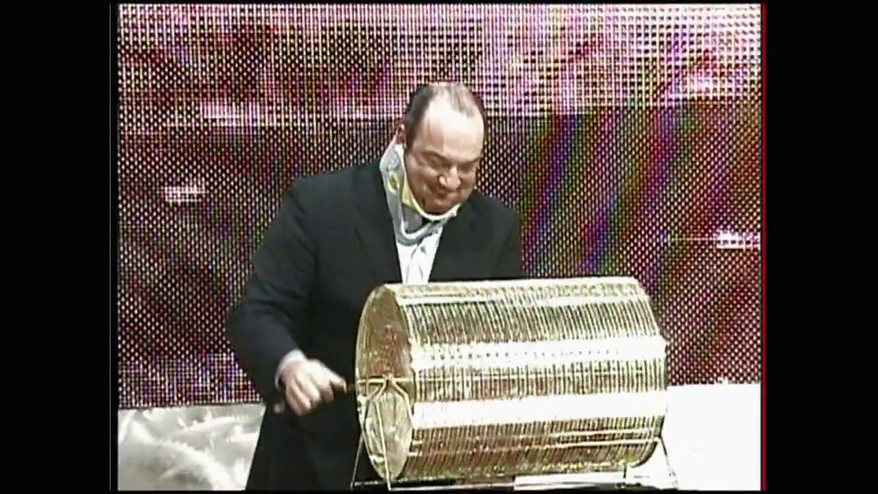 A frame from WWE Raw (March 29, 2004) at 0:26. A man in a black suit and white shirt stands at a table, examining a large woven basket or container. The backdrop features a colorful pixelated LED screen typical of wrestling event production.