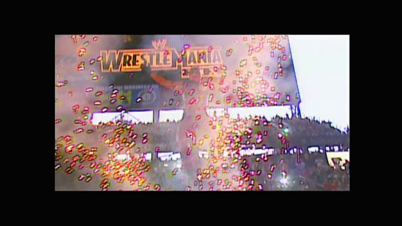 A frame from WWE Raw (March 31, 2003) at 0:48. The WrestleMania logo appears prominently at the top of the screen with gold and red confetti falling across the frame. The view shows an outdoor wrestling venue with a mountainous backdrop visible behind the arena structure and crowd seating.