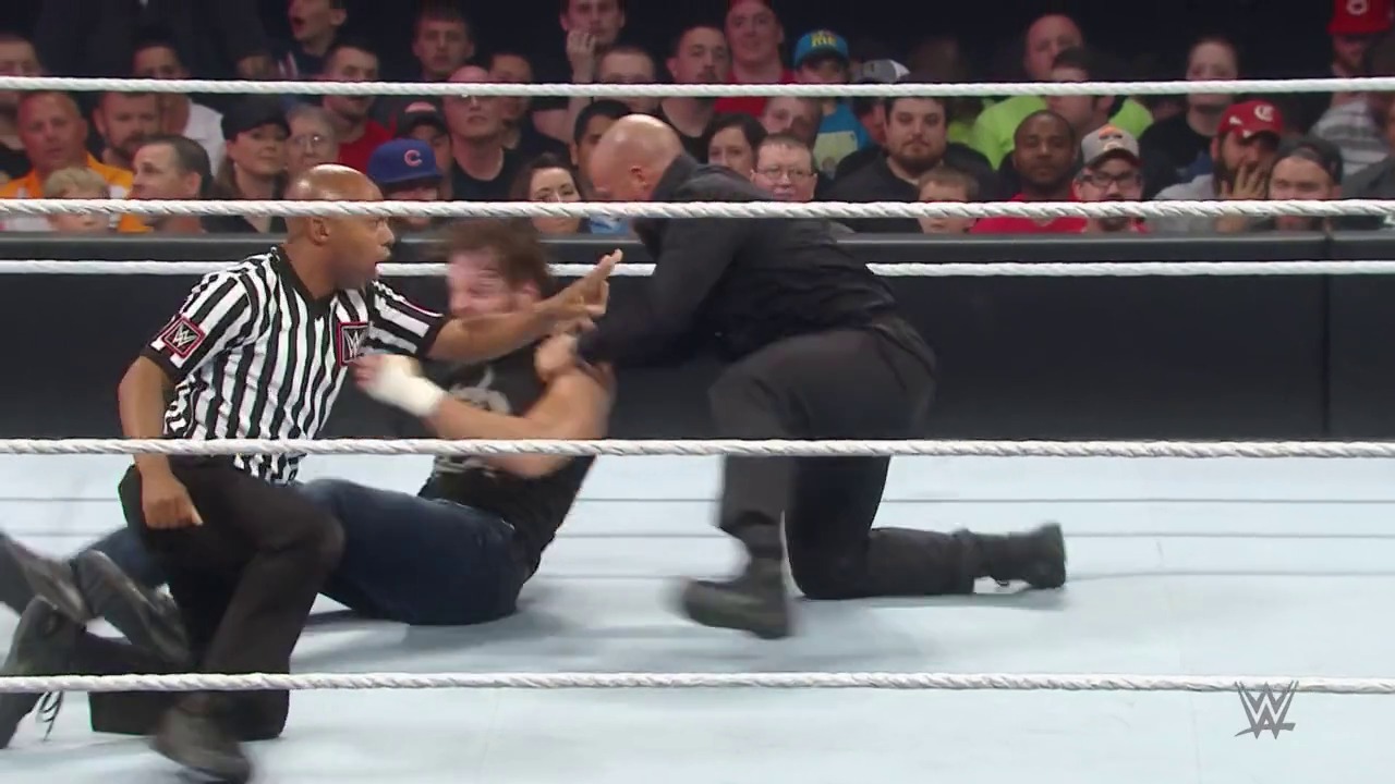 A frame from WWE Raw (May 11, 2015) at 20:01. A WWE referee in a striped shirt kneels on the white wrestling ring, gesturing toward a wrestler in black clothing. A crowd of spectators fills the arena seating in the background.