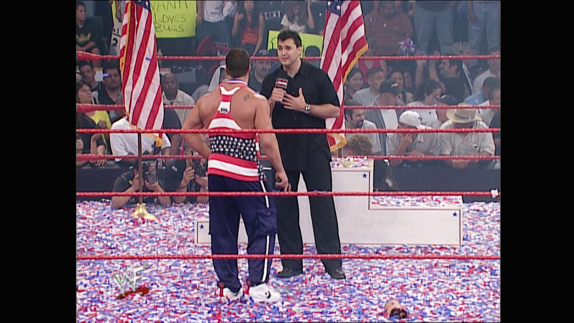 A frame from WWE Raw (May 21, 2001) at 48:11. A WWE wrestler in patriotic attire stands in the ring with a microphone-wielding announcer, surrounded by American flags and confetti. The crowd fills the background behind the red ropes of the wrestling ring.