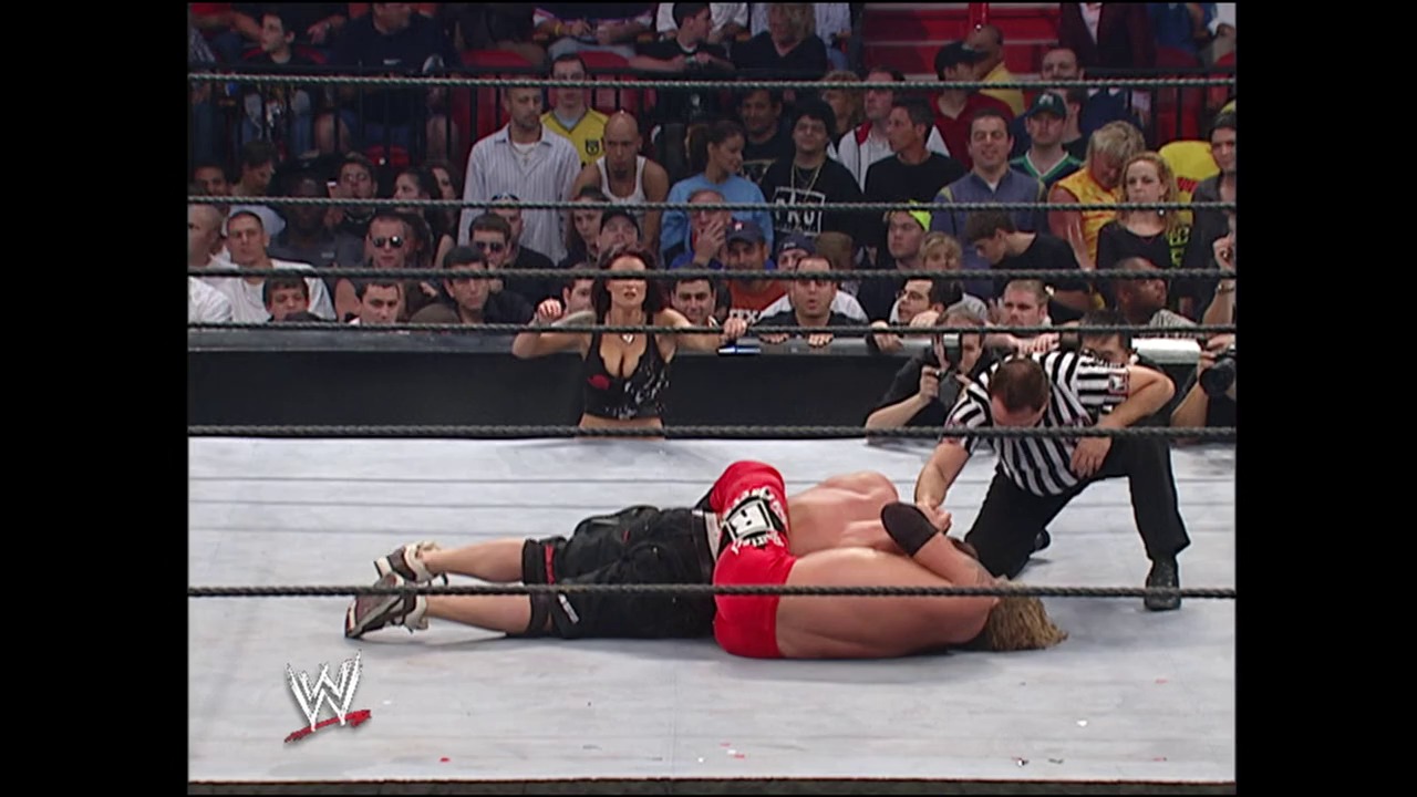 A frame from WWE Royal Rumble (2006) at 2:21:03. A wrestler in red and black attire lies on the mat wearing a red bandana, while a referee in striped shirt stands nearby. The crowd fills the arena stands behind the ring ropes in what appears to be a WWE Royal Rumble match.