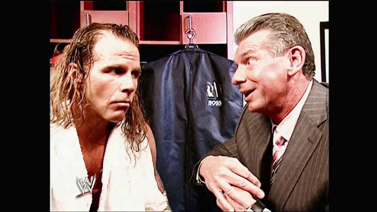 A frame from WWE Royal Rumble (2006) at 46:48. A WWE wrestler with long, wet hair in a white shirt stands facing a man in a brown suit and tie during what appears to be a backstage interview segment. A dark jacket hangs on a hook in the background.