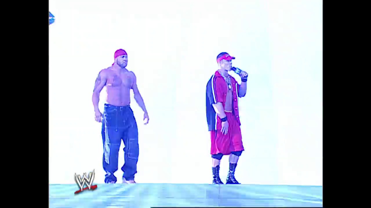 A frame from WWE SmackDown (January 30, 2003) at 12:13. Two wrestlers stand in the WWE ring during a SmackDown event. One shirtless wrestler wears blue cargo pants, while the other holds a microphone and wears a pink/magenta outfit with a baseball cap, ready to address the crowd.