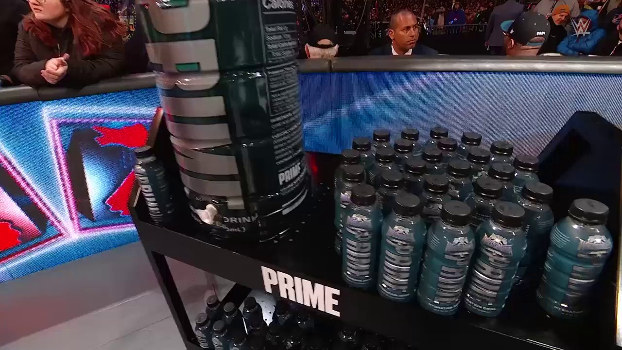 A frame from WWE WrestleMania 40 Saturday (2024) at 2:27:34. A PRIME energy drink display stands ringside at WWE WrestleMania 40, featuring multiple bottles arranged on a black promotional stand with the brand logo, while the illuminated ring and crowd are visible in the background.