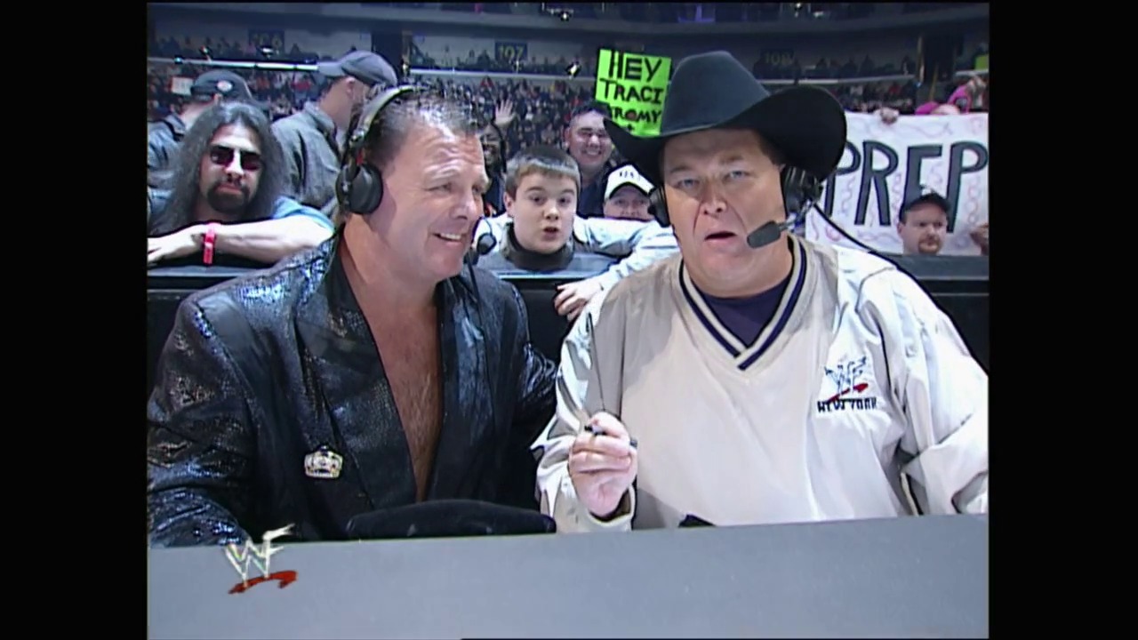 A frame from Royal Rumble (2001) at 48:44. Two men in wrestling attire sit at a commentary table during a WWE event, with one wearing a dark sequined jacket and the other in a white shirt and black cowboy hat. A crowd fills the arena in the background with various signs visible.
