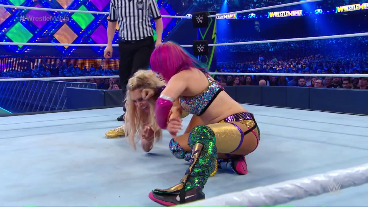 A frame from WrestleMania (2018) at 43:28. # WrestleMania Wrestling Match

A wrestler with magenta hair wearing colorful sequined attire dominates an opponent on the white wrestling ring canvas. A referee in striped shirt oversees the match while the crowd and illuminated WrestleMania graphics are visible in the background.