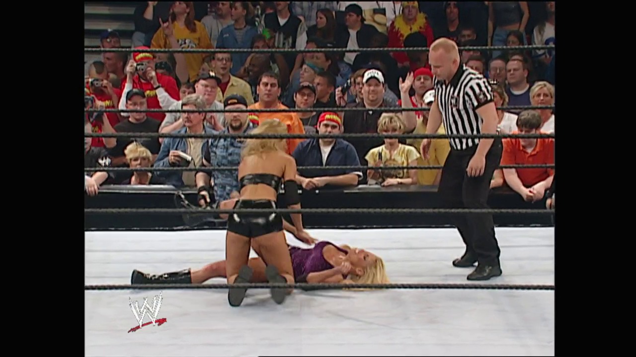 A frame from WWE Judgment Day (2002) at 23:54. A WWE wrestler in black attire stands over a fallen opponent in purple on the white canvas, while a referee in striped shirt officiates. The crowd fills the arena seats in the background, with the WWE logo visible in the lower left corner.