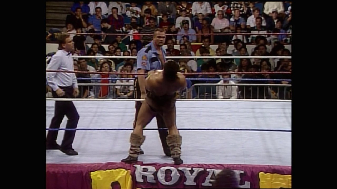 A frame from WWE Royal Rumble 1991 (1991) at 39:50. A wrestler in tan/beige attire stands in a WWE Royal Rumble ring with a confident stance, while officials and spectators fill the arena in the background. The iconic "ROYAL" text is visible on the ring apron padding.