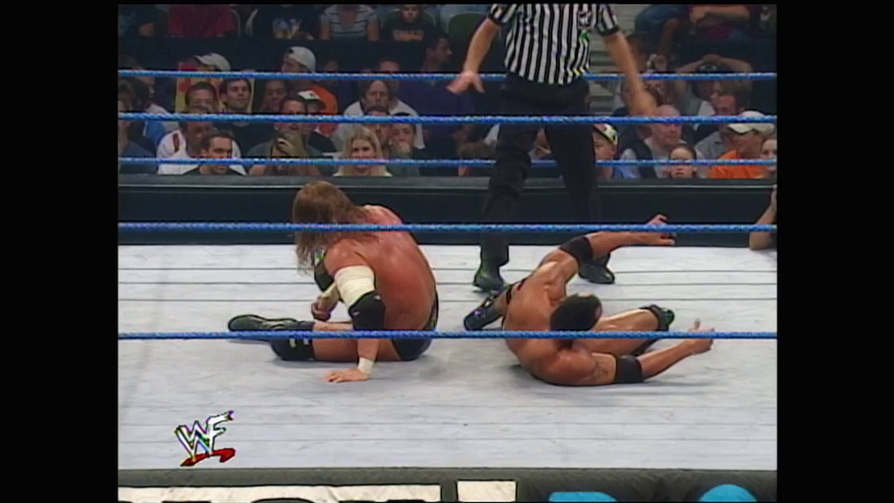 A frame from WWE SmackDown (August 17, 2000) at 1:26:57. Two wrestlers are down on the white mat of a WWE ring, with one appearing to be in pain or exhausted. A referee in a striped shirt stands nearby as the crowd watches from the blue-roped ring perimeter, with the WWF logo visible in the corner.