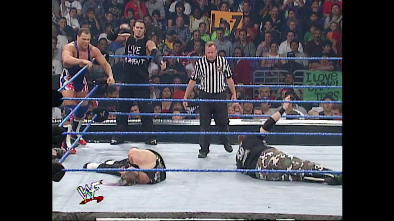 A frame from WWE SmackDown (August 9, 2001) at 1:16:37. A WWE SmackDown match in progress with two wrestlers in the ring, one standing and one on the canvas. A referee in striped shirt oversees the action while a large crowd watches from the stands, with the WWF logo visible in the corner.