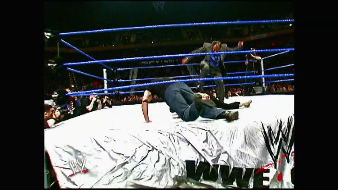 A frame from Royal Rumble (2004) at 23:54. A wrestler lies on the white mat inside a WWE ring with blue ropes, while another competitor stands nearby. The crowd is visible in the background, and the WWE logo is displayed on the ring apron.