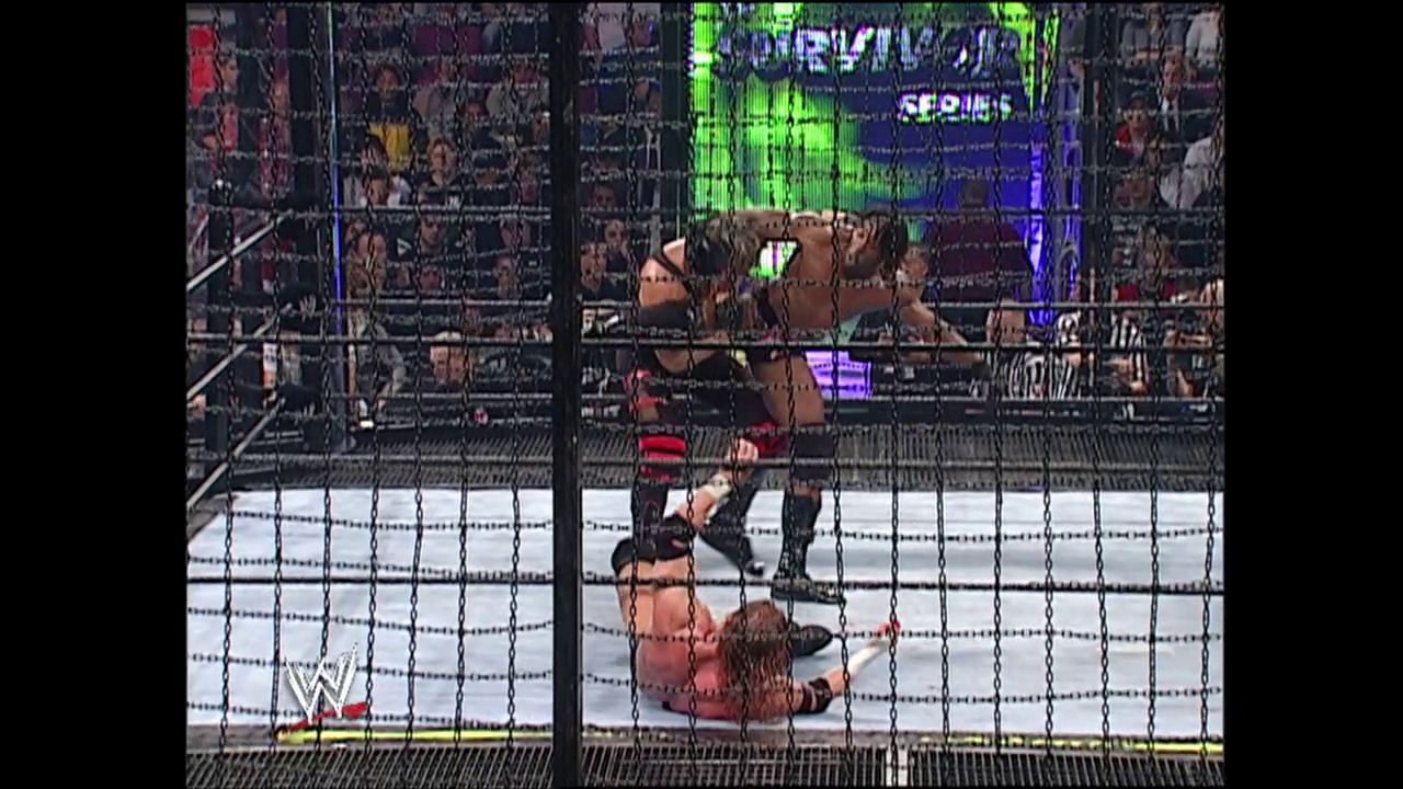 A frame from Survivor Series (2002) at 2:19:34. A wrestler lies on the steel cage floor while another stands nearby in a WWE Survivor Series match. The crowd fills the arena in the background as green and blue graphics display on the overhead screen.