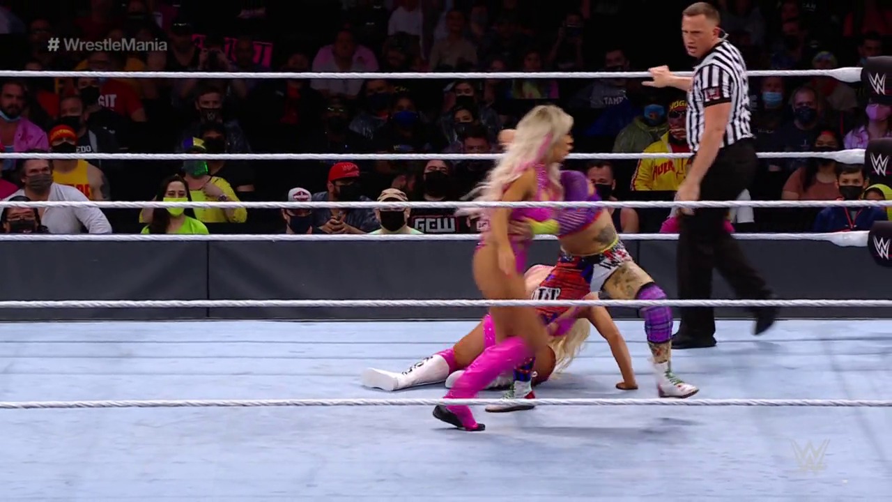 A frame from WrestleMania 37 (2021) at 54:18. A blonde female wrestler in bright pink and purple attire is on the mat during a WrestleMania match, with a referee in a striped shirt overseeing the action. The crowd is visible in the background behind the white-roped WWE ring.