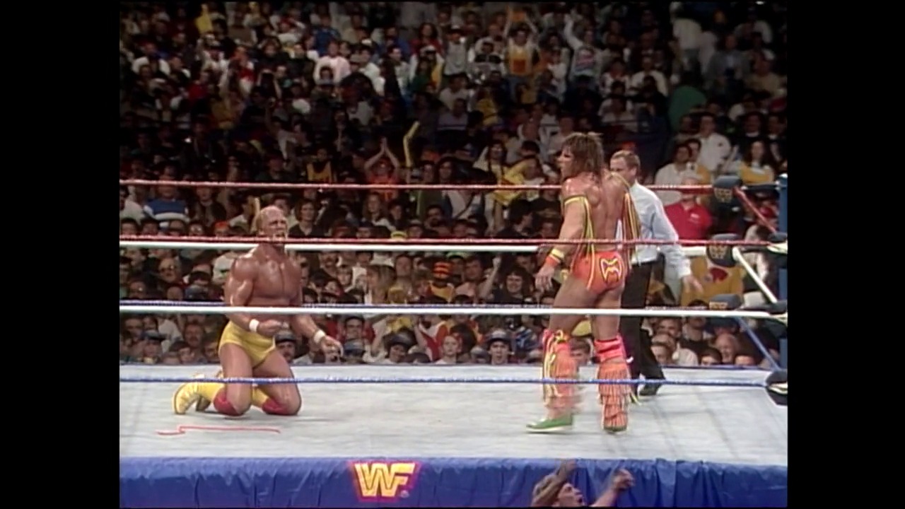 A frame from WrestleMania VI (1990) at 3:17:44. A WWE wrestler in yellow and red attire kneels in the ring while another wrestler stands nearby, with a packed crowd visible in the background and the WWF logo displayed on the ring apron.