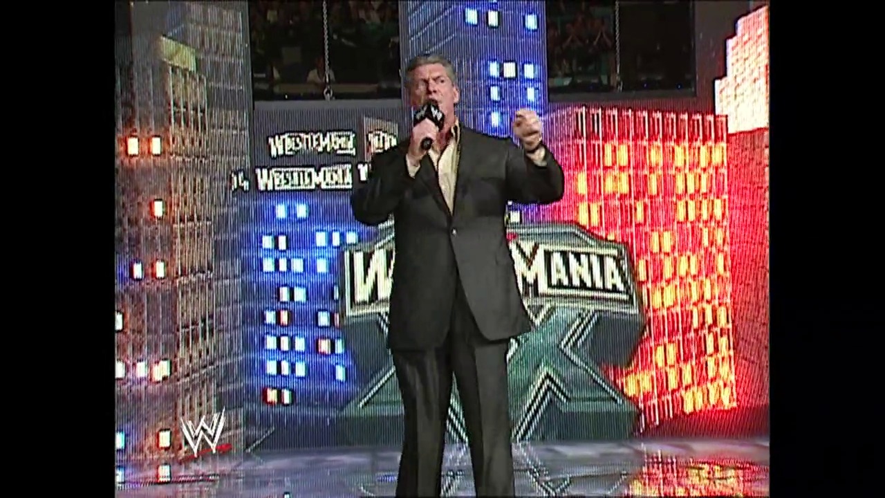 A frame from WrestleMania XX (2004) at 2:34:20. A man in a dark suit speaks into a microphone on stage at WrestleMania XX, flanked by illuminated building-shaped set pieces in blue and red with the WrestleMania logo visible in the background.