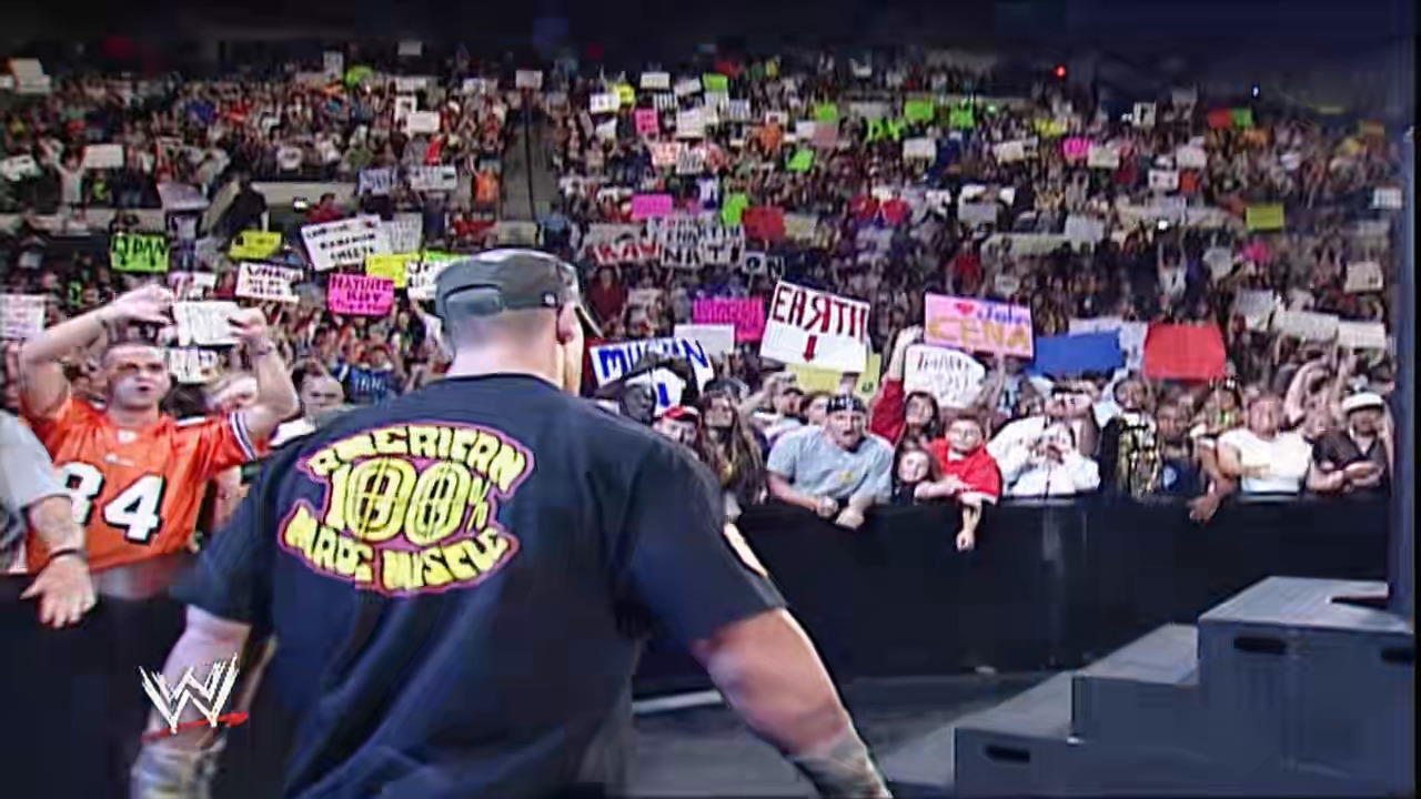 A frame from WWE Raw (May 14, 2007) at 0:58. A WWE wrestler wearing a navy "American Badass" shirt stands in the ring facing a massive crowd holding colorful signs and placards. The arena is packed with enthusiastic fans, with the WWE logo visible in the lower left corner.