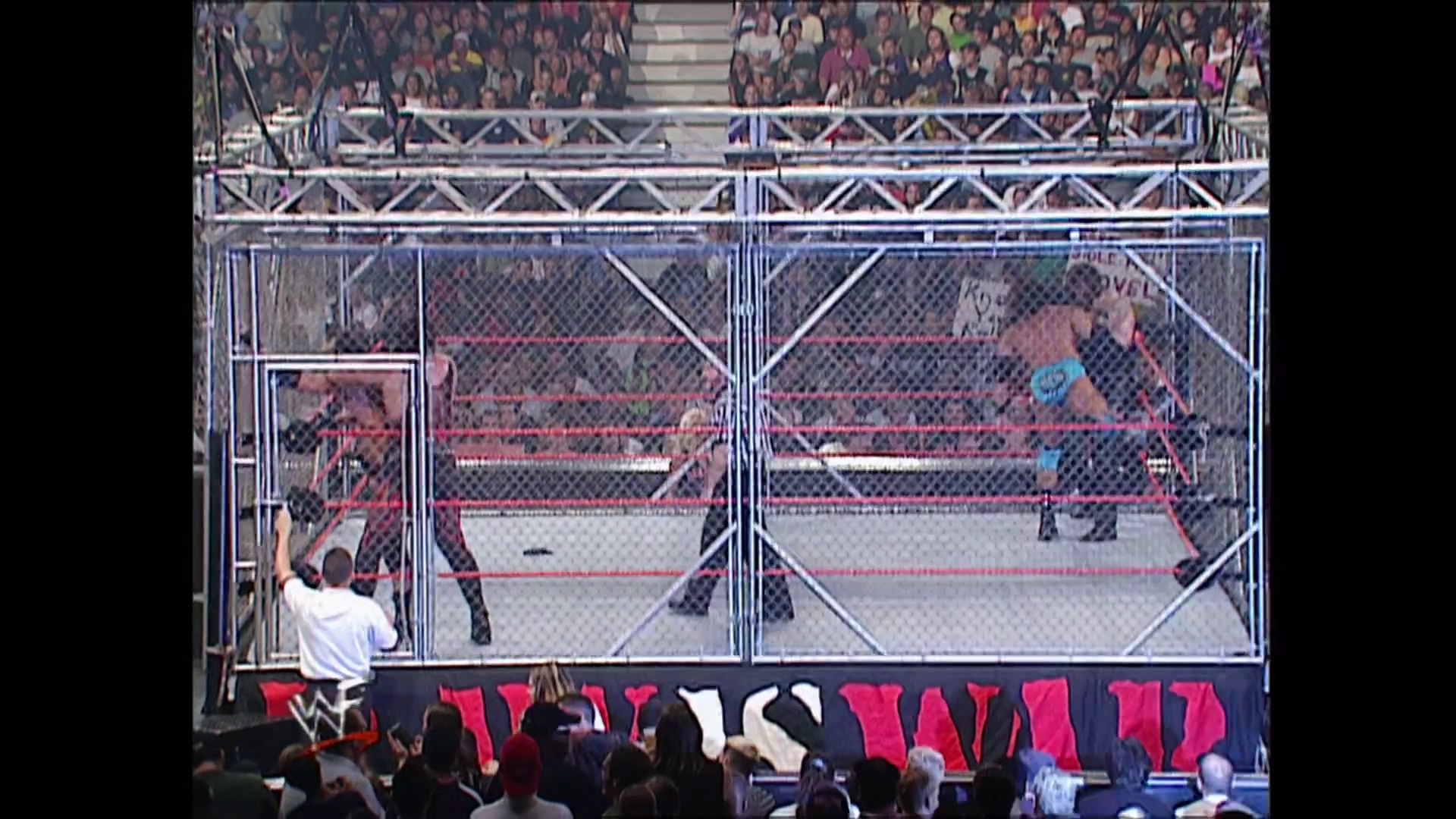 A frame from WWE Raw (August 13, 2001) at 41:42. A wrestler in a white shirt stands outside a steel cage match setup with red ropes and chain-link fencing, while opponents are visible inside the ring. Crowds fill the arena stands above the branded WWE barrier.