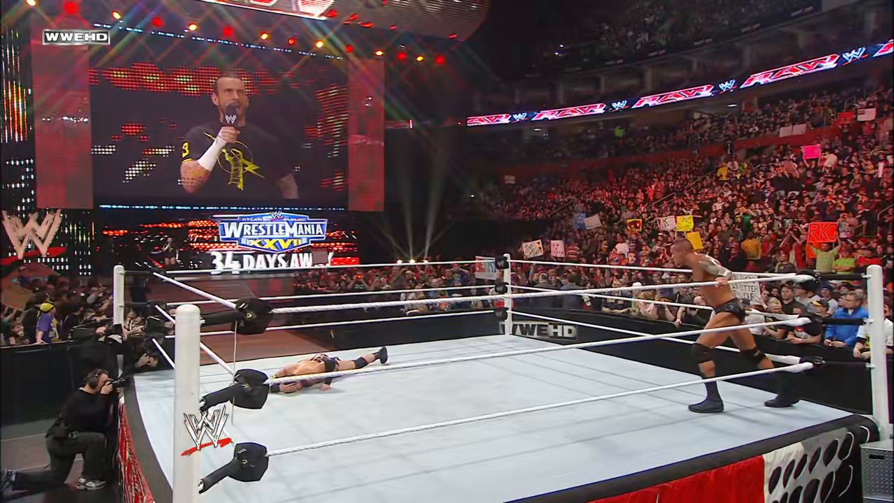 A frame from WWE Raw (February 28, 2011) at 41:43. A WWE Raw wrestling match in progress, with one wrestler lying on the mat while another stands in the ring. The arena is packed with enthusiastic fans, and large video screens display the action, with "WrestleMania XXVII" and "34 Days Away" graphics visible.