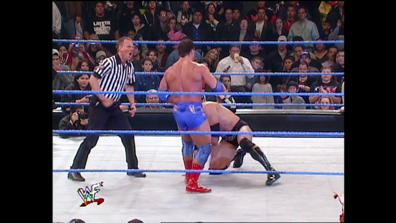 A frame from WWE SmackDown (January 4, 2001) at 39:11. A WWE wrestler in blue and red attire executes a move on an opponent in the ring while a referee in a striped shirt supervises. The crowd watches intently from the stands surrounding the illuminated blue-roped wrestling ring.