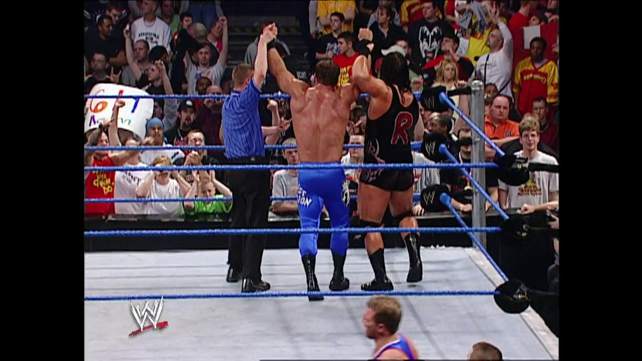 A frame from WWE SmackDown (March 20, 2003) at 1:11:45. A WWE wrestler in blue attire celebrates with raised arms in the ring while opponents lie on the mat, with an energetic crowd visible in the background and the WWE logo displayed at ringside.