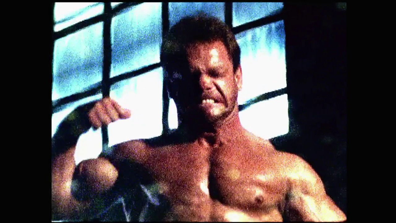 A frame from Survivor Series (2003) at 0:19. A muscular male wrestler stands shirtless in a wrestling ring, posed in a confrontational stance with his fist raised. Large illuminated windows are visible in the background, creating dramatic lighting for what appears to be a professional wrestling broadcast.