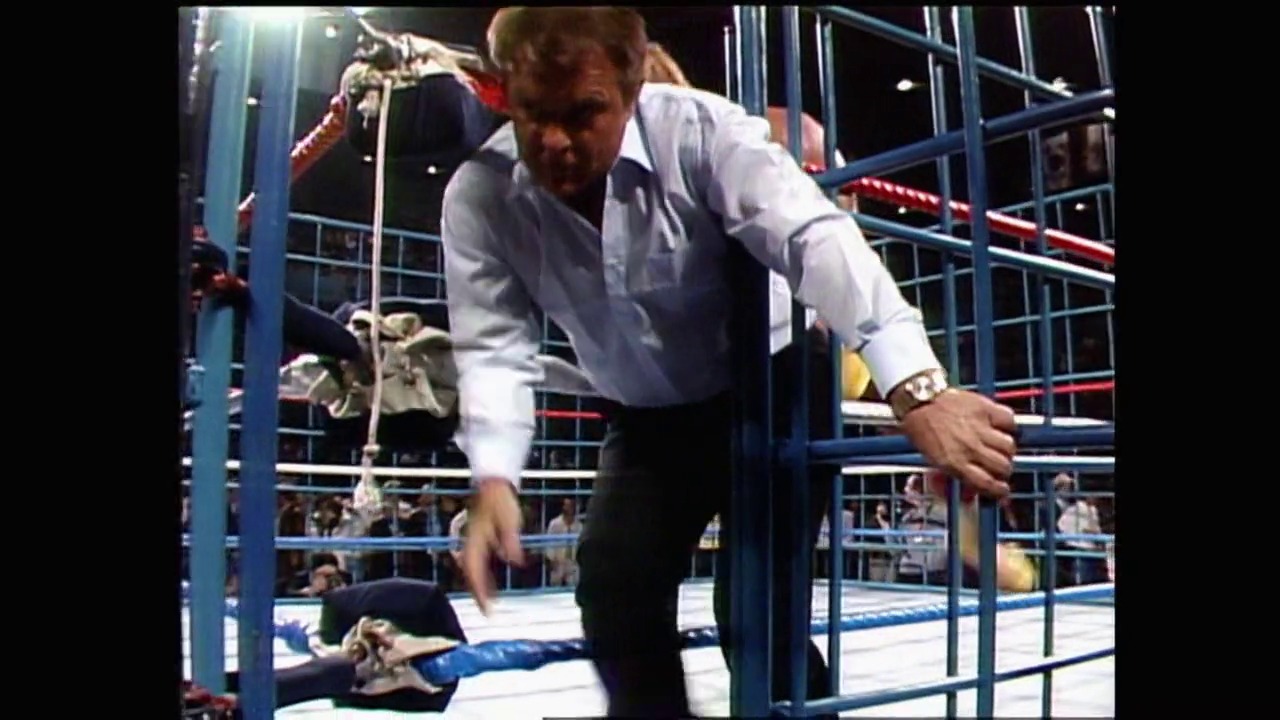 A frame from WrestleMania 2 (1986) at 3:01:04. A referee in a white shirt leans over a fallen wrestler on the blue mat inside a wrestling ring, while spectators watch from the stands during a professional wrestling match.