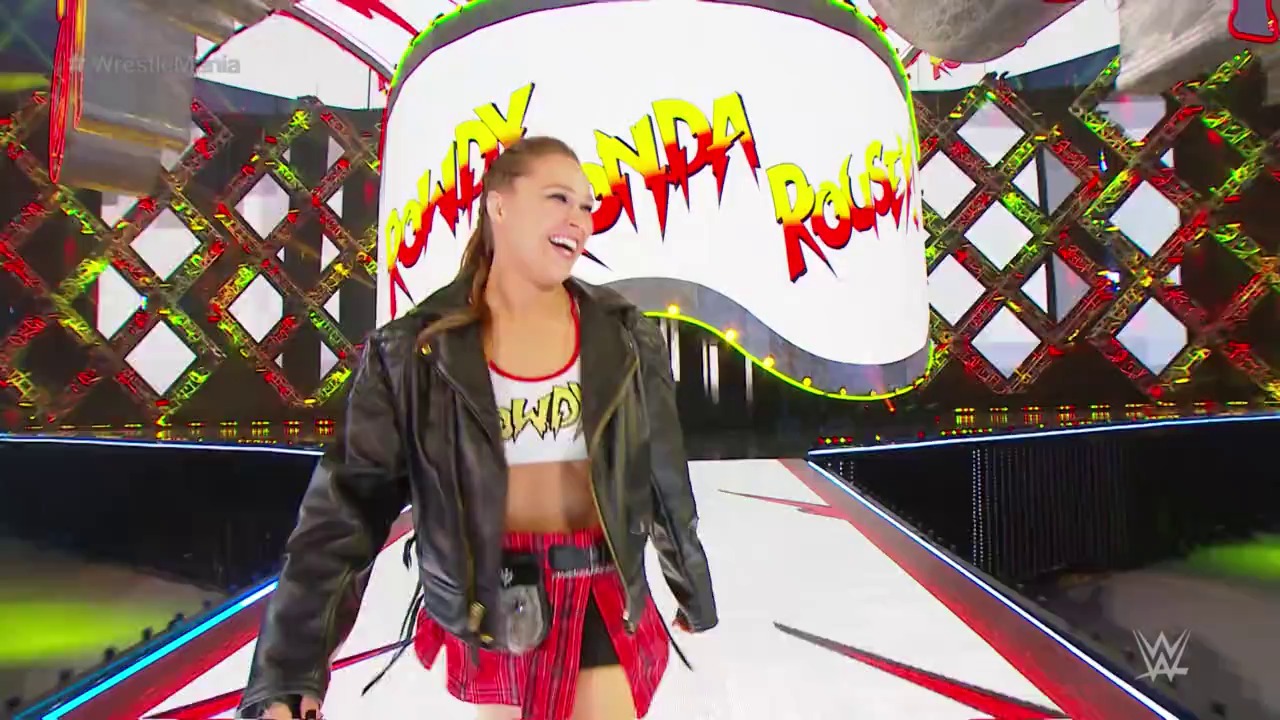 A frame from WrestleMania (2018) at 1:27:15. A female wrestler stands on the WrestleMania ring entrance area, smiling while wearing a black leather jacket over a white crop top and red skirt. Vibrant neon graphics with "RONDA" text and colorful decorative elements frame the stage behind her.