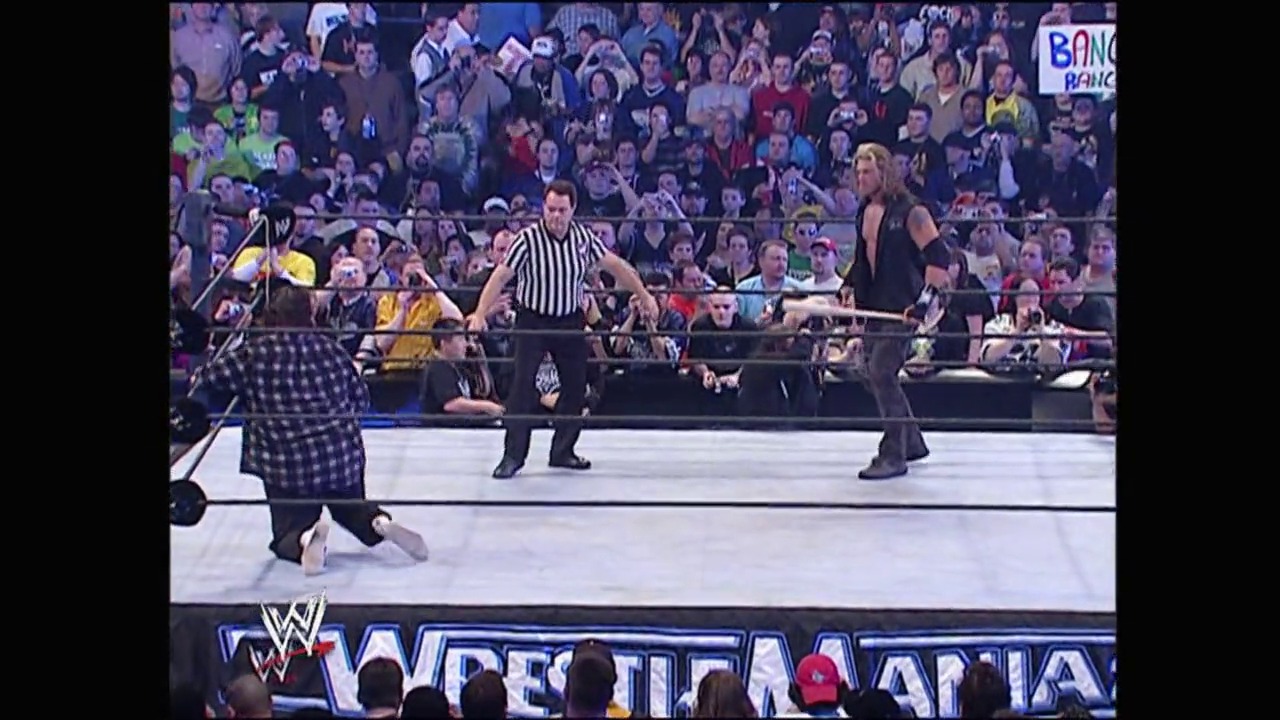 A frame from WrestleMania 22 (2006) at 1:06:50. A WWE wrestling match is underway at WrestleMania 22, with a referee in striped shirt overseeing two wrestlers in the ring. The packed crowd fills the arena stands while the WWE logo and event graphics are visible at the bottom of the screen.