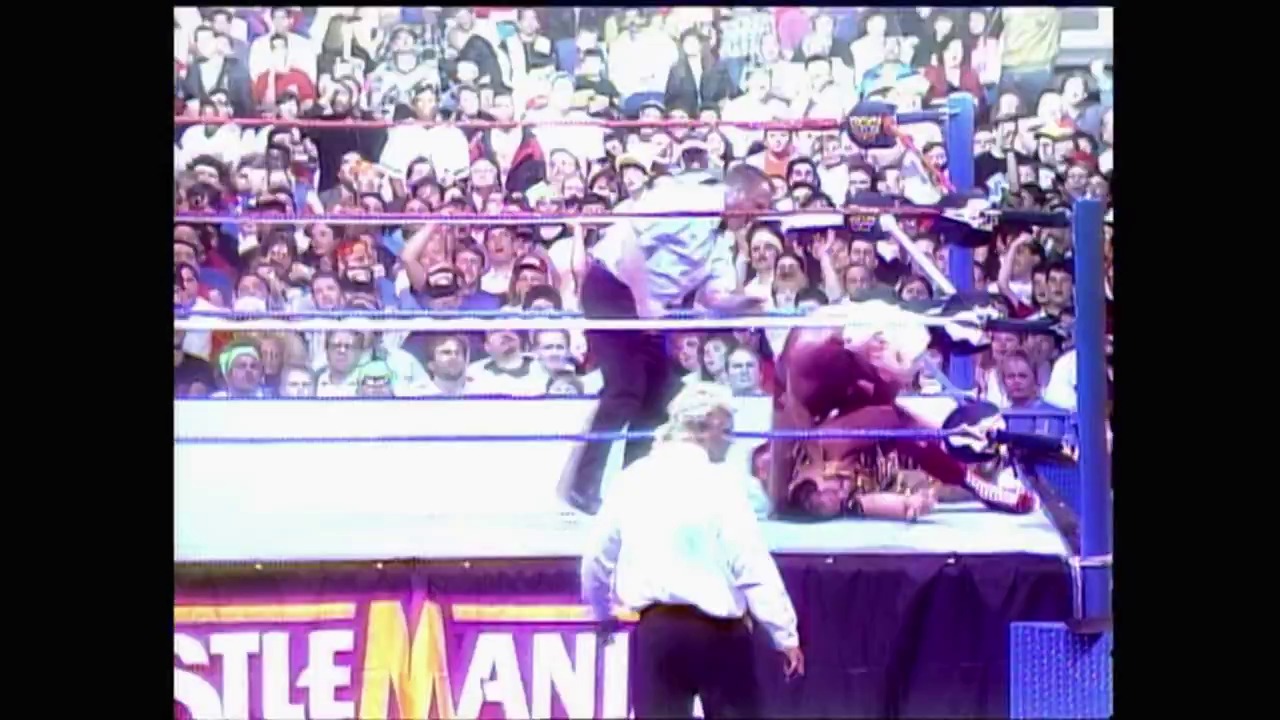 A frame from WrestleMania VIII (1992) at 1:31:39. A wrestler in white shirt and dark pants stands in the ring at WrestleMania VIII, with a packed crowd visible in the bleachers behind him. The event branding is visible at the bottom of the frame, and the wrestling ring is surrounded by blue ropes and barriers.