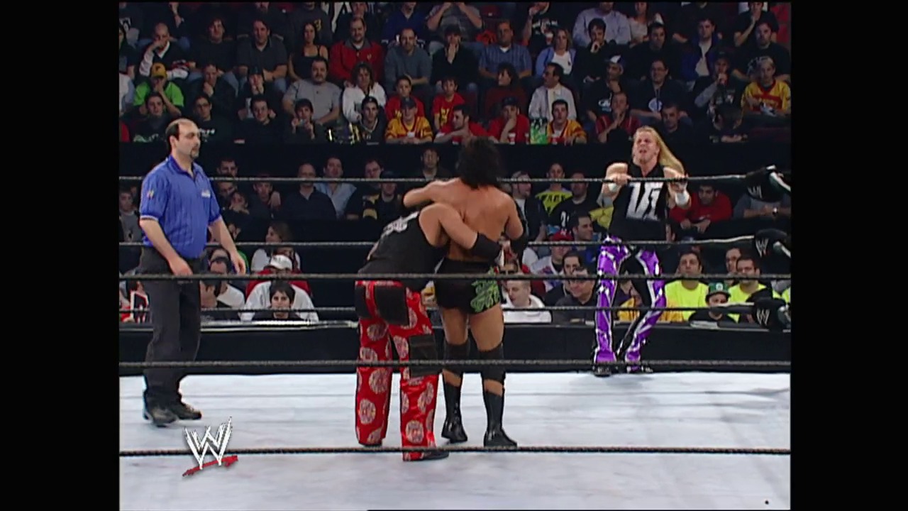 A frame from WWE No Way Out (2003) at 43:58. A WWE wrestling match in progress with two wrestlers in the ring, one shirtless in black pants and another in purple and black attire. A referee in blue oversees the match while the crowd watches from the stands, with the WWE logo visible at the ringside.