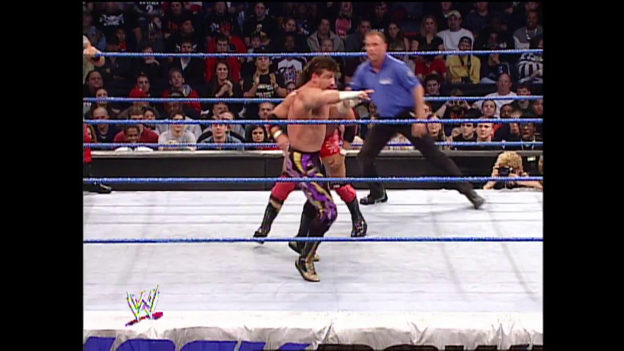 A frame from WWE Raw (December 12, 2002) at 22:52. A shirtless WWE wrestler in red and purple attire executes a move in the blue-roped ring while a referee in blue watches. The crowd fills the arena stands in the background, with the WWE logo visible in the lower left corner.