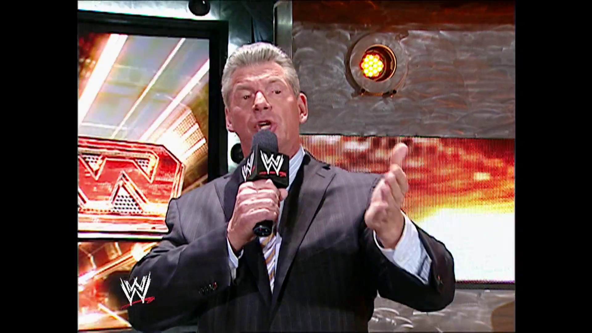 A frame from WWE Raw (November 20, 2006) at 13:18. A WWE official in a dark suit and tie speaks into a microphone on stage, gesturing with his hand. Behind him are illuminated WWE graphics and screens displaying red and gold branding typical of WWE Raw programming.