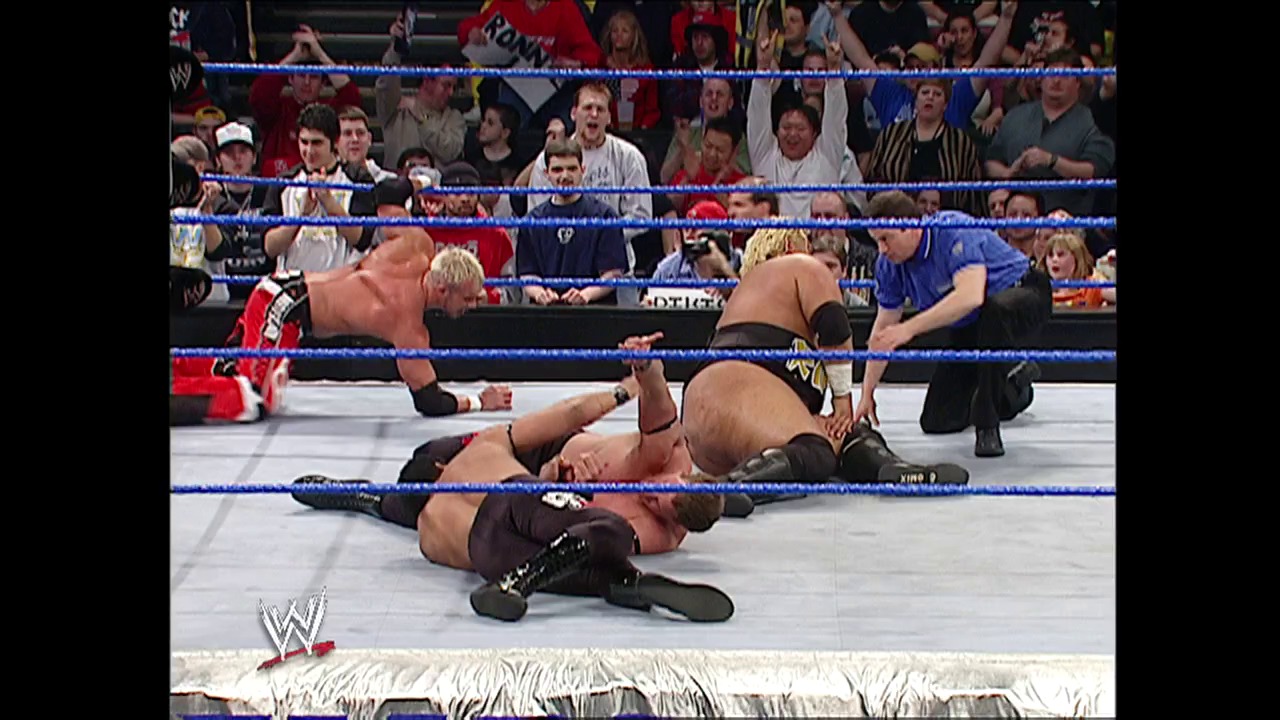 A frame from WWE SmackDown (February 26, 2004) at 24:51. A WWE wrestler lies on the mat during a SmackDown match, with another competitor nearby and a referee attending to the action. The blue-roped ring is surrounded by an engaged crowd, with the WWE logo visible in the corner.