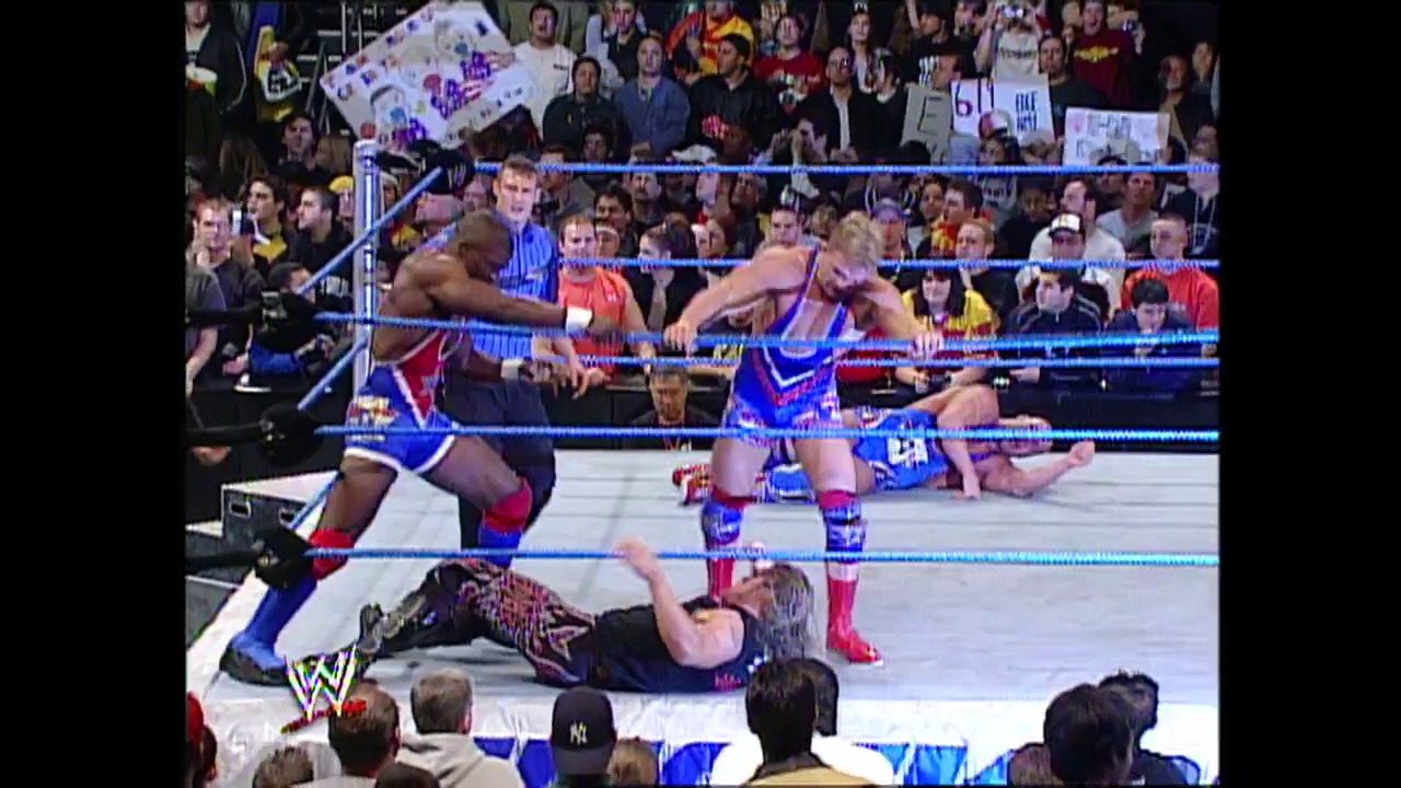 A frame from WWE SmackDown (February 6, 2003) at 1:23:38. A WWE wrestler lies on the mat while two opponents stand over him in the blue-roped ring, surrounded by an enthusiastic crowd. The WWE logo is visible in the lower left corner of the broadcast.