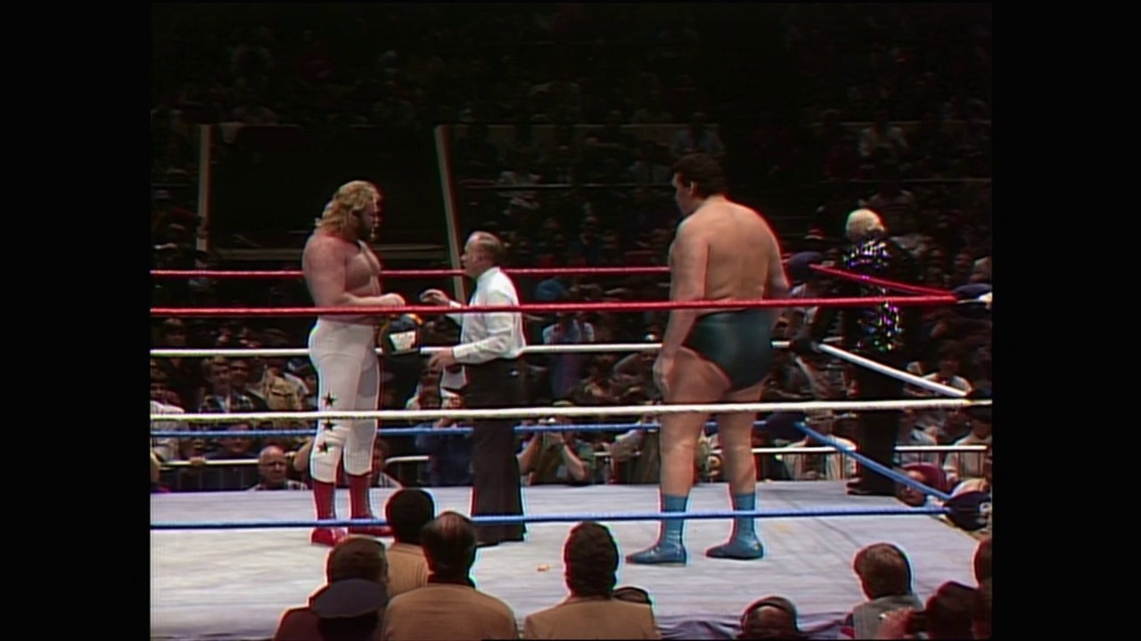 A frame from WrestleMania I (1985) at 1:18:16. Two wrestlers face off in a professional wrestling ring at WrestleMania I, with a referee in a white shirt between them. The crowd fills the arena around the blue and red roped ring, captured in vintage 1985 footage quality.