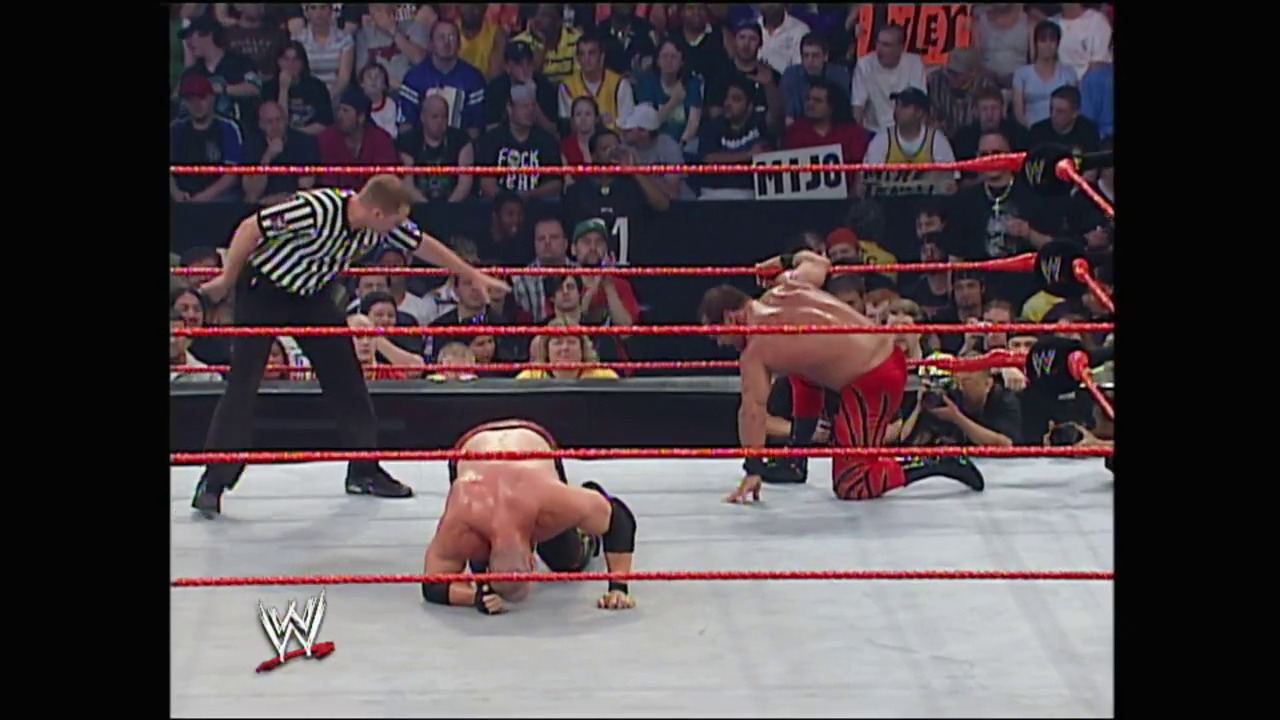 A frame from WWE Bad Blood (2004) at 1:38:09. A WWE wrestler in red and black attire is on the mat in a prone position, while a referee in a striped shirt stands nearby in the red-roped wrestling ring. The crowd fills the arena stands in the background, with the WWE logo visible at the bottom of the frame.