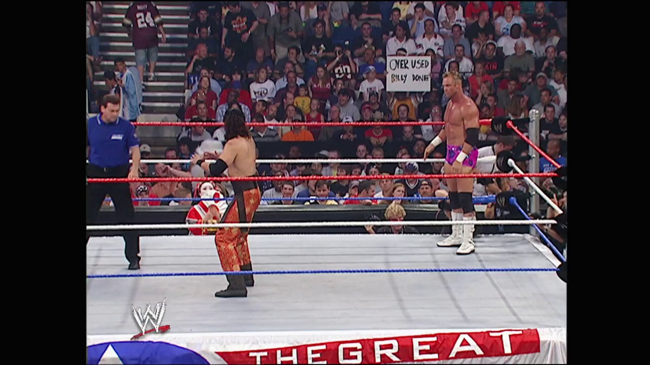 A frame from WWE Great American Bash (2004) at 1:10:41. A WWE wrestling match is underway in a packed arena, with three wrestlers in and around the ring—one shirtless wrestler in purple wrist tape stands opposite another competitor, while a third in red and gold attire is positioned in the foreground. The crowd fills the stands behind the red and blue roped ring, with visible signage and the WWE logo displayed at the bottom of the frame.