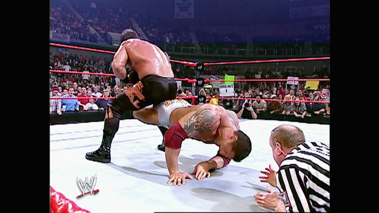 A frame from WWE Raw (December 13, 2004) at 53:38. A WWE wrestler performs a move on an opponent in the ring during Raw, with the referee nearby monitoring the match. The crowd is visible in the background of the arena, and the red ropes and WWE branding are prominently displayed.