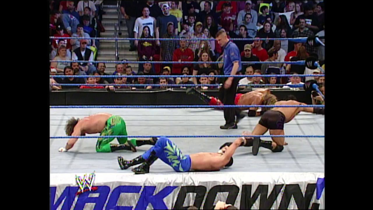 A frame from WWE Raw (December 26, 2002) at 37:02. Multiple wrestlers are down on the WWE SmackDown ring canvas during an intense match, with a referee standing over them and a packed crowd visible in the bleachers behind the blue ropes.